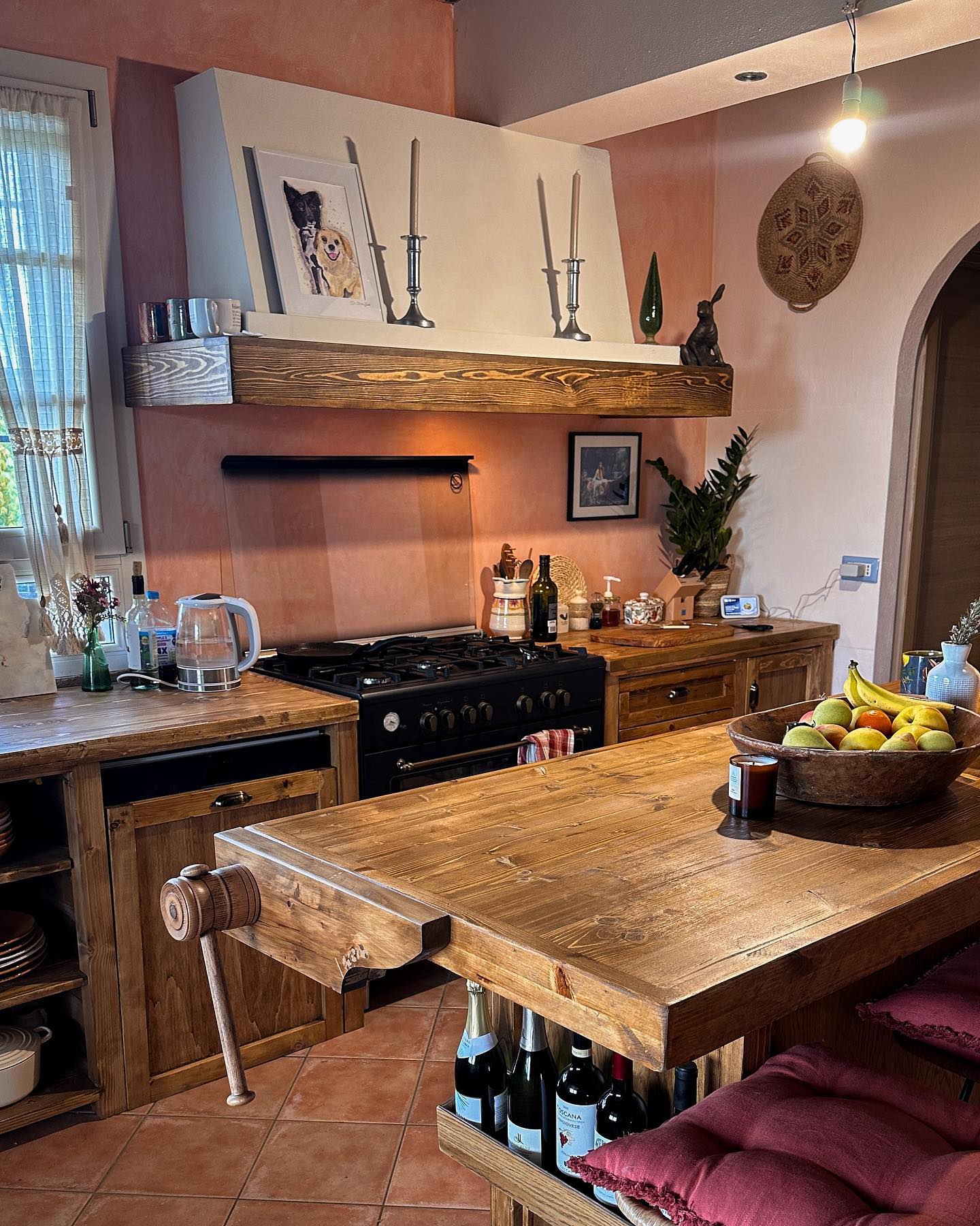 Lived-in Tuscan kitchen with terracotta pink plaster walls, vintage pine carpenter workbench used as an island complete with wooden vice, rustic pine cabinets, black range, wood mantel beam with silver candlesticks, and terracotta tile floor