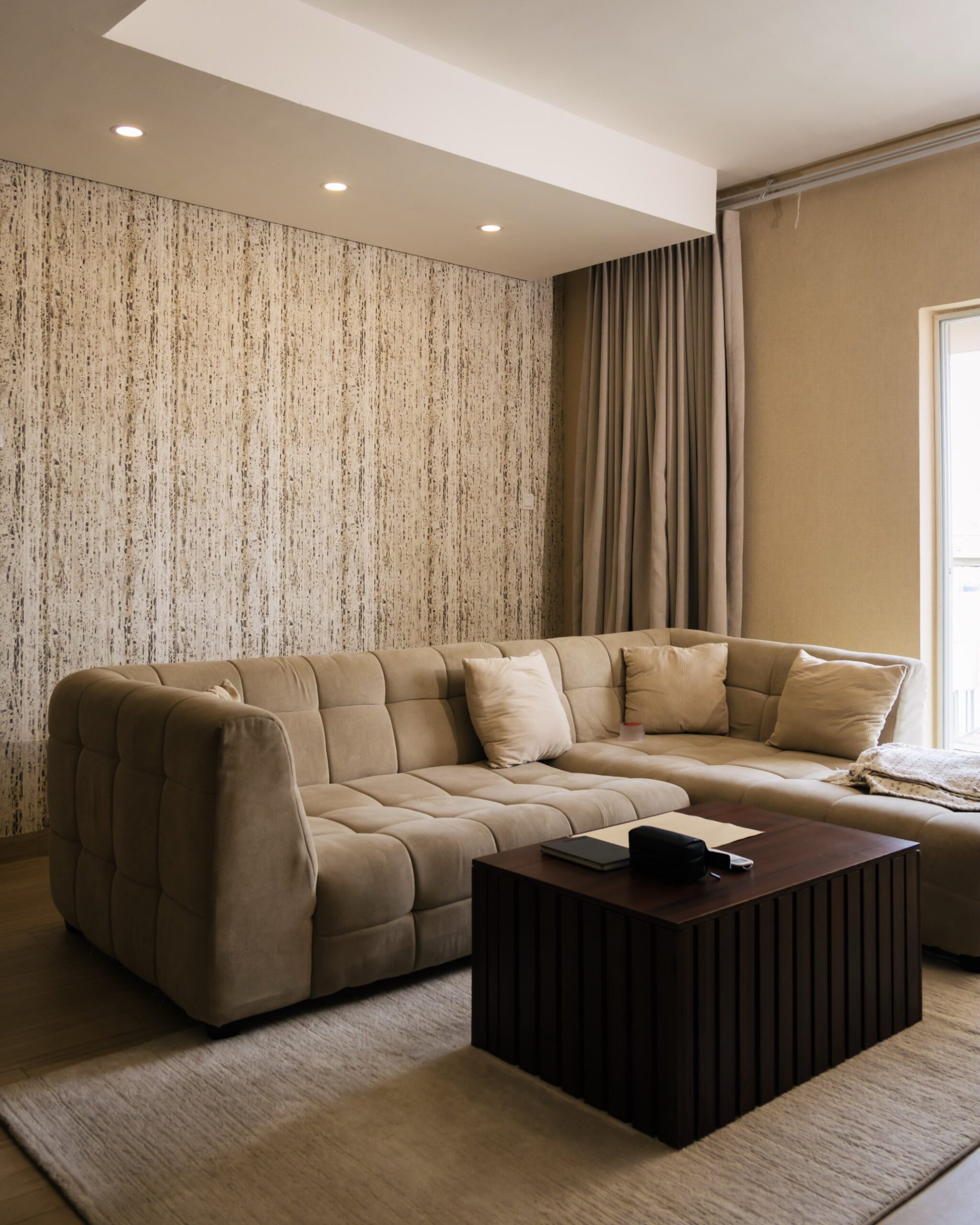Mottled vertical beige and cream textured wallpaper behind a plush tufted beige sectional with cream pillows, a dark wood slatted square coffee table, and warm neutral curtains