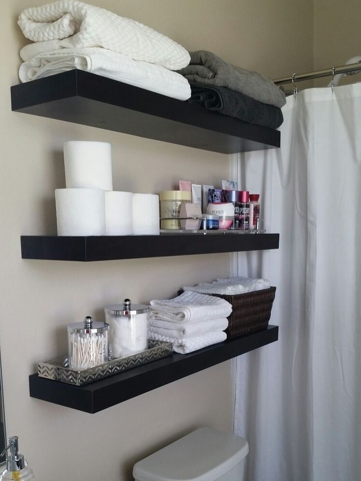 Three dark floating shelves mounted on a cream wall above a white toilet, holding stacked towels, backup toilet paper, lotions, and apothecary jars
