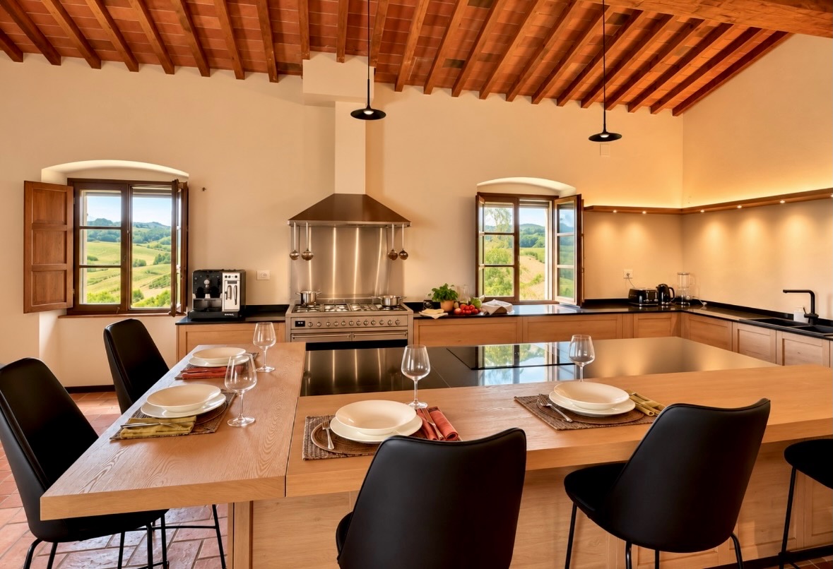 Modern Tuscan kitchen with pitched timber beam ceiling and terracotta tile insets, light oak cabinets, black counters, stainless pro range with pot rail, twin arched windows with wood shutters framing hillside views, and a large island with built-in cooktop plus an oak dining extension
