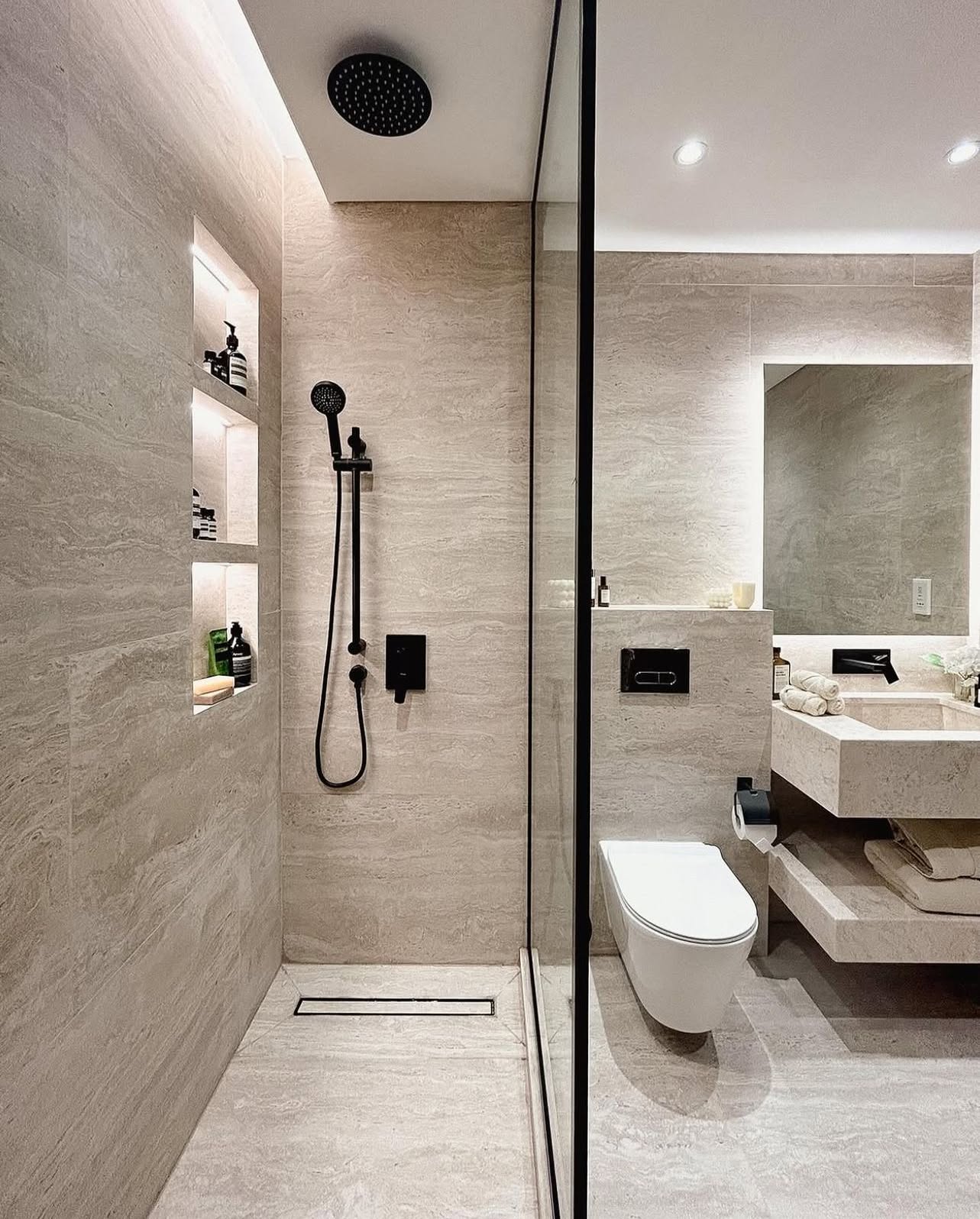 Small travertine shower bathroom with floor-to-ceiling cream stone tiles, black ceiling-mounted rain shower head, black handheld shower on a slide bar, three stacked backlit niches, floating stone vanity with an undermount sink and tall backlit mirror