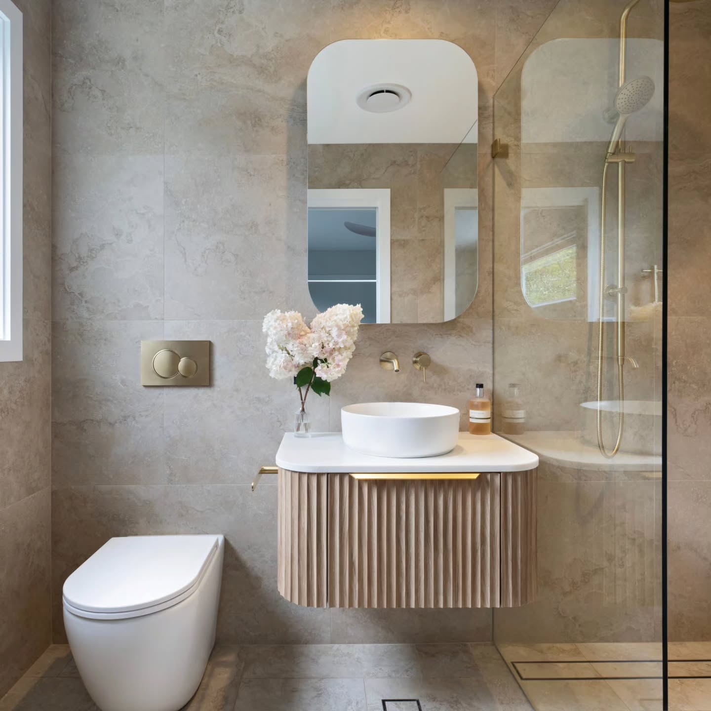 Small travertine bathroom with a floating fluted wood vanity, white vessel sink, brass wall-mounted tap, long slim mirror, wall-hung toilet with a brass flush plate and a walk-in shower with brass fixtures behind glass