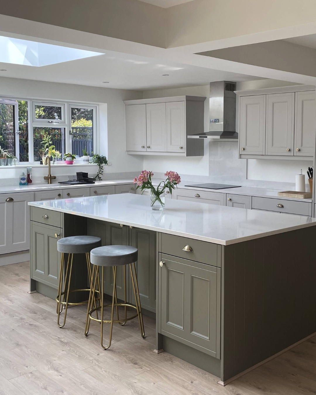 Two-tone kitchen with light greige perimeter shaker cabinets, a sage green island with a white quartz top, and two gray velvet backless barstools on gold hairpin legs