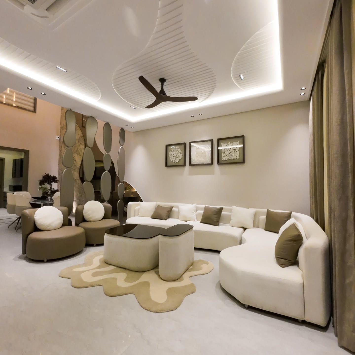 Long white curved sectional sofa with brown and cream pillows and two round taupe swivel armchairs with white round pillows, facing a sculptural cream coffee table set on a wavy cream and tan rug, with a dramatic undulating white tray ceiling with LED cove lighting, a dark three-blade ceiling fan, a triptych of textured artworks on the back wall, and a tall metallic mirror sculpture wall on the left