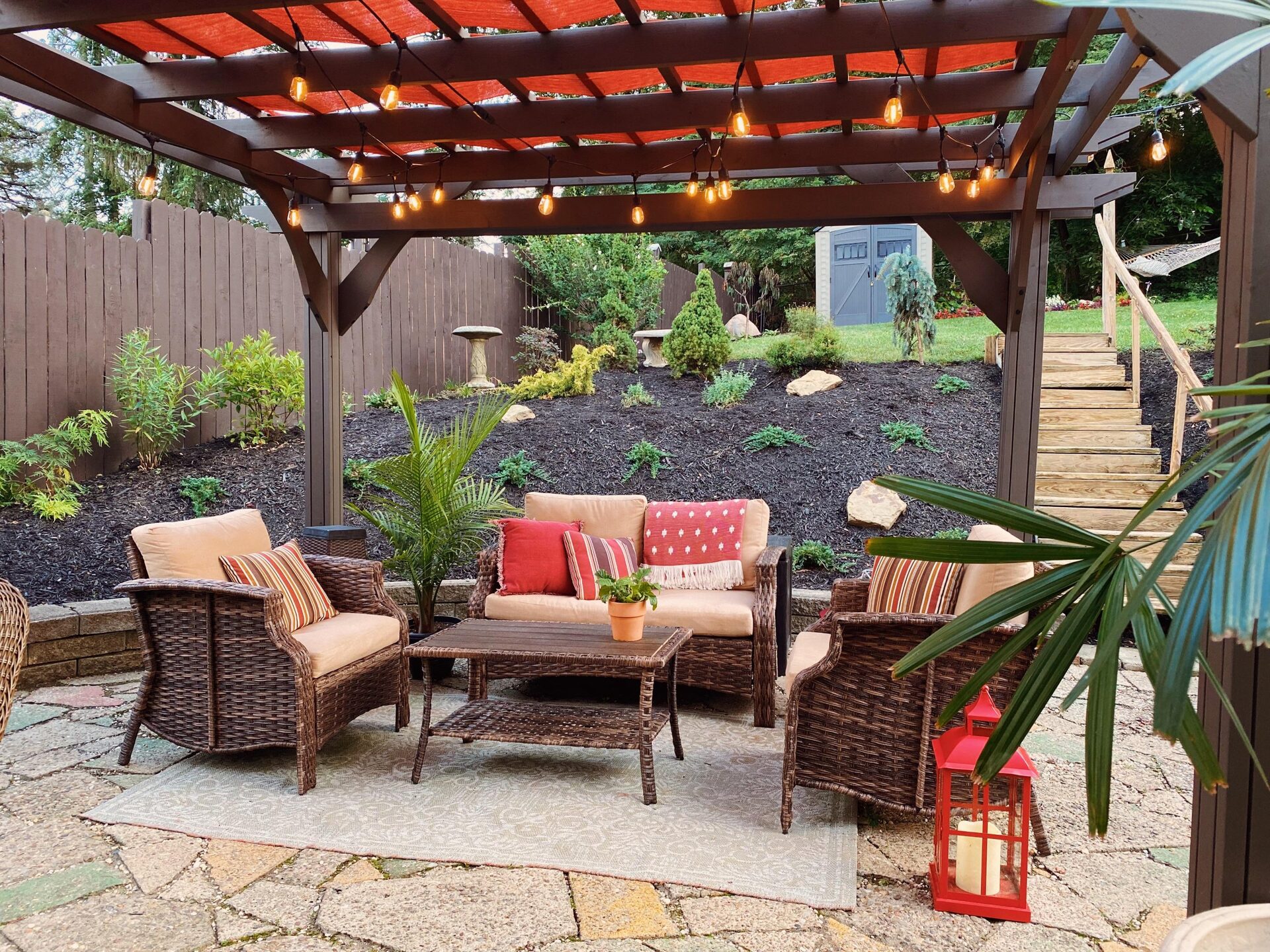 Upper backyard lawn with a gray storage shed, red coleus flower bed, hammock strung between trees, a tall white birdhouse on a pole, and the red shade sail of the pergola in the lower corner.