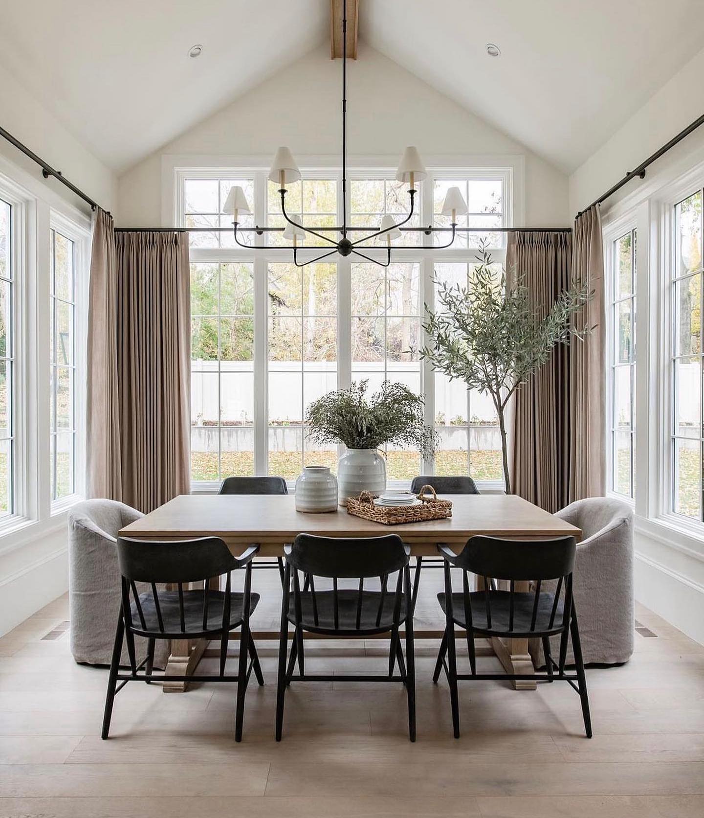 Modern farmhouse dining nook with vaulted white ceiling, tall windows on three sides, wood trestle table, black spindle chairs on the long sides, and soft gray slipcovered chairs on each