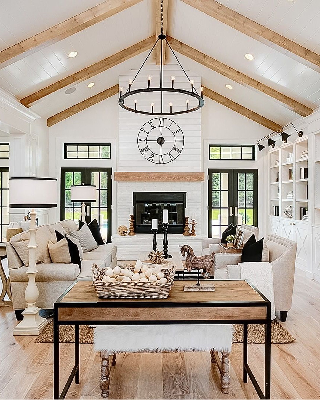 Modern farmhouse living room with vaulted ceiling, exposed wood beams, round black chandelier, and oversized clock over a white brick fireplace