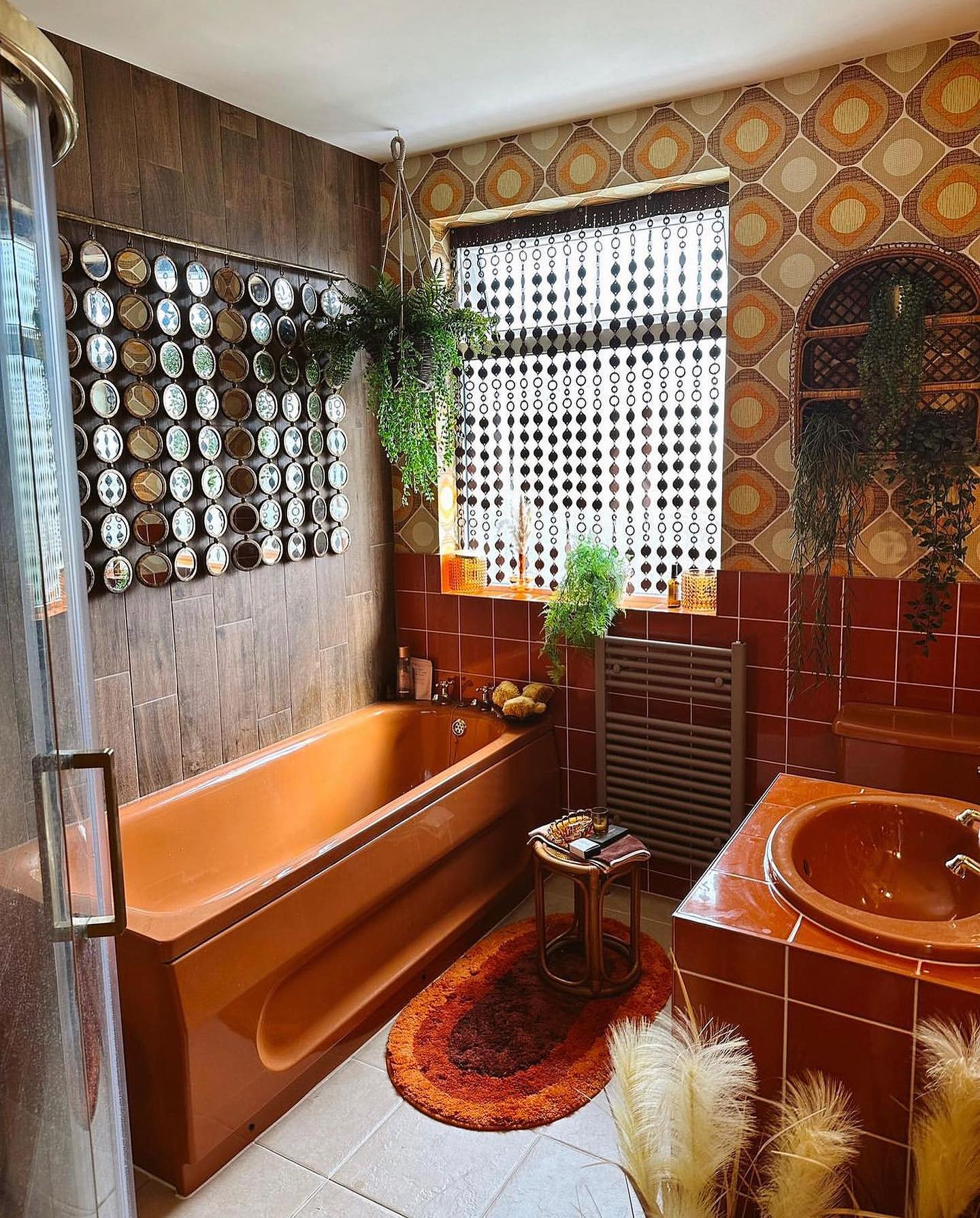 Bold 1970s bathroom with burnt orange tub and sink, brown and orange geometric wallpaper, wall of small round mirrors, hanging ferns, and bead curtain over window