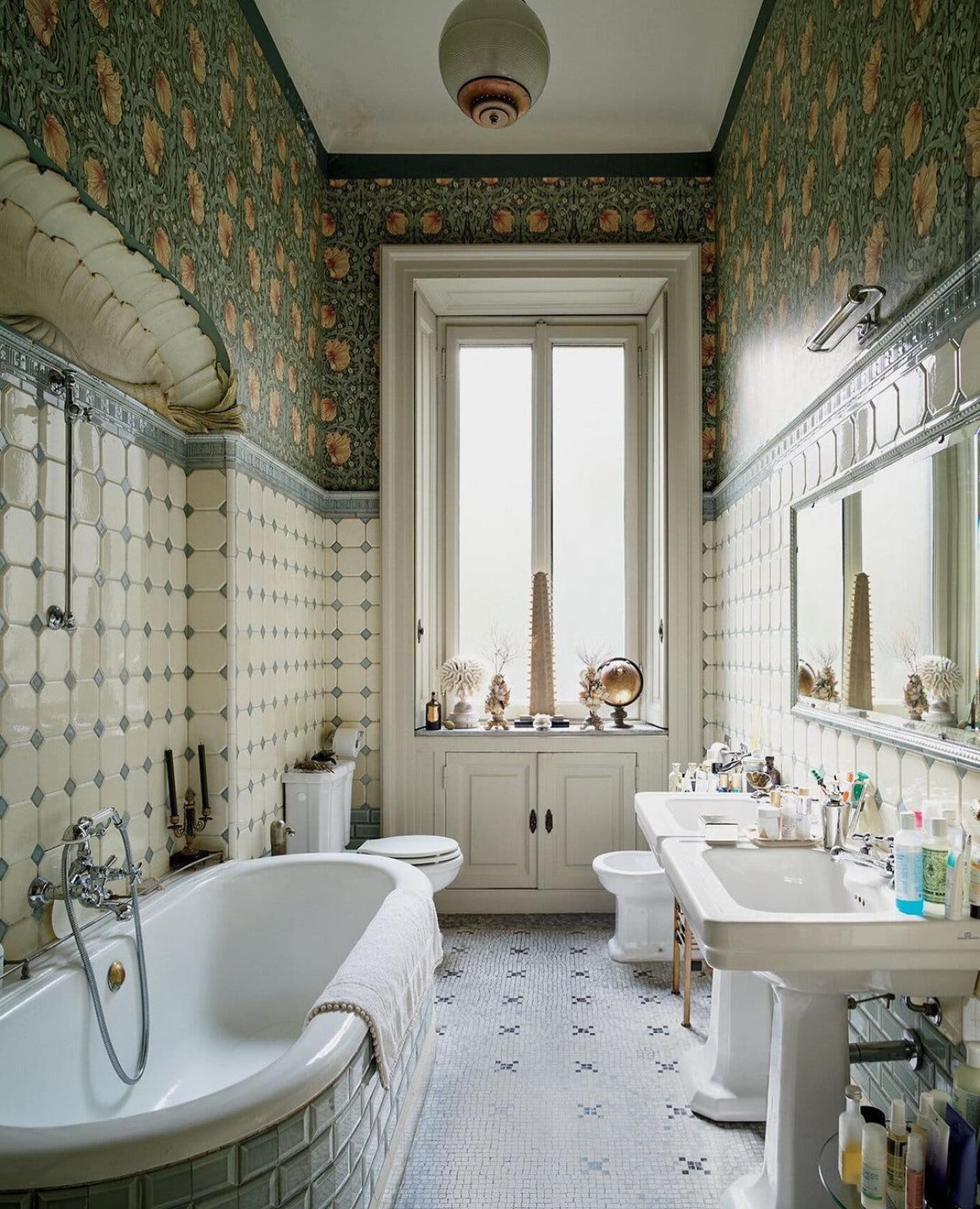 European vintage bathroom with Morris floral wallpaper, white octagon tile wainscot, pedestal sink, soft curved freestanding tub, and small mosaic floor pattern