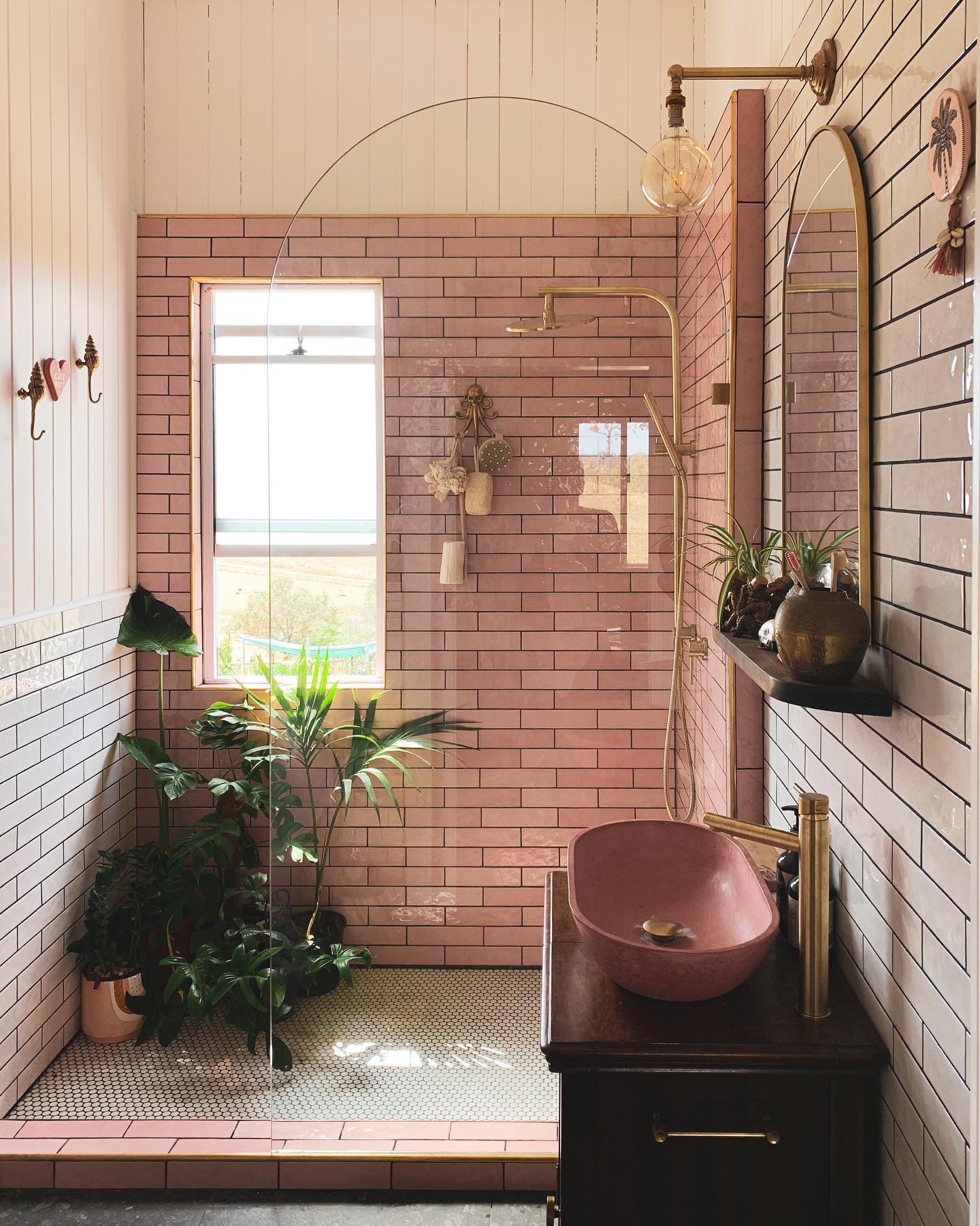 Pink subway tile shower with brass rain showerhead, pink concrete vessel sink, dark wood vanity, white subway tile wall, and large tropical plants in the corner