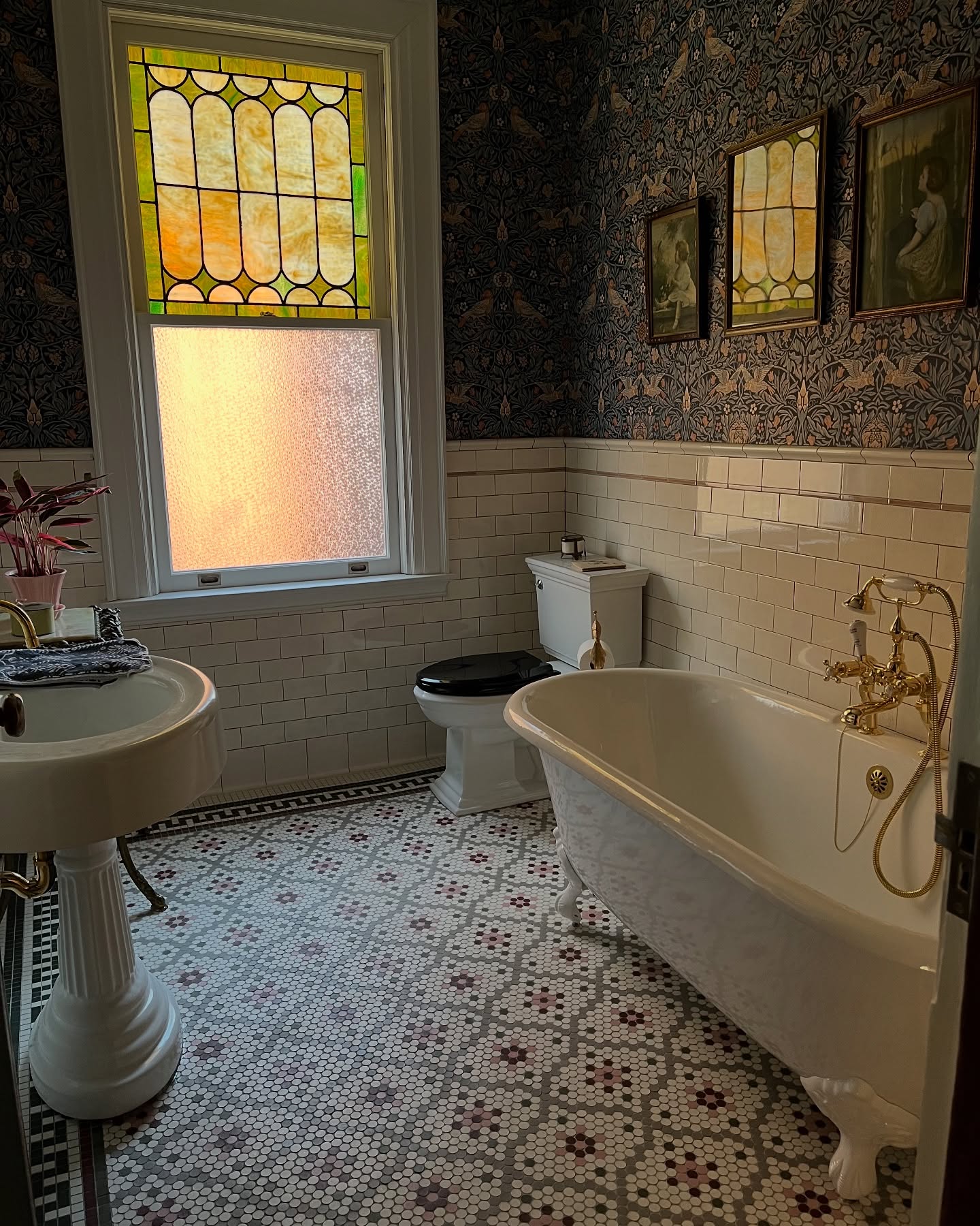 Victorian bathroom with dark Morris bird wallpaper, amber stained glass window, white clawfoot tub with gold fixtures, and penny tile floor with flower pattern