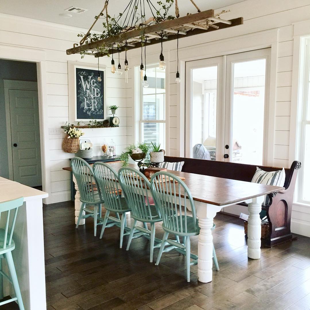 Modern farmhouse dining room with a vintage wooden ladder hung horizontally as a chandelier with Edison bulbs and greenery woven through, sage green Windsor dining chairs, white farm table, and a dark wood church pew bench