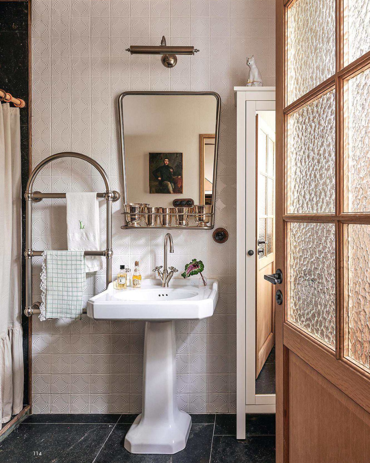 Vintage powder room with white embossed fan-pattern wallpaper, pedestal sink, polished nickel towel warmer, framed portrait reflected in mirror, and dark stone floor