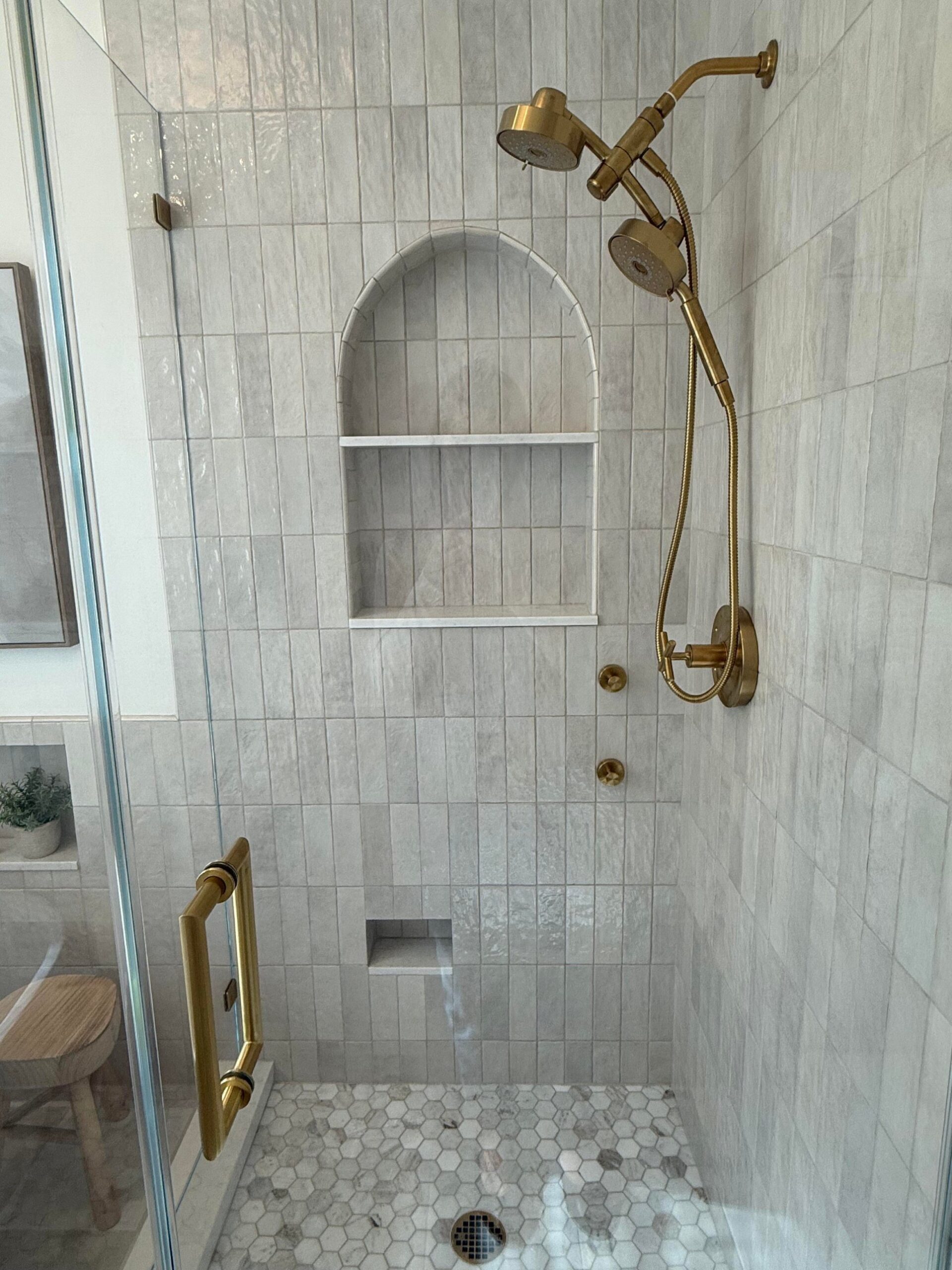 Walk-in shower with pale vertical tile, a tall arched niche with a shelf, a wall-mount brass shower head, a brass handheld shower on a slide bar, brass controls, a marble hex tile floor, and a glass door with a long brass handle.