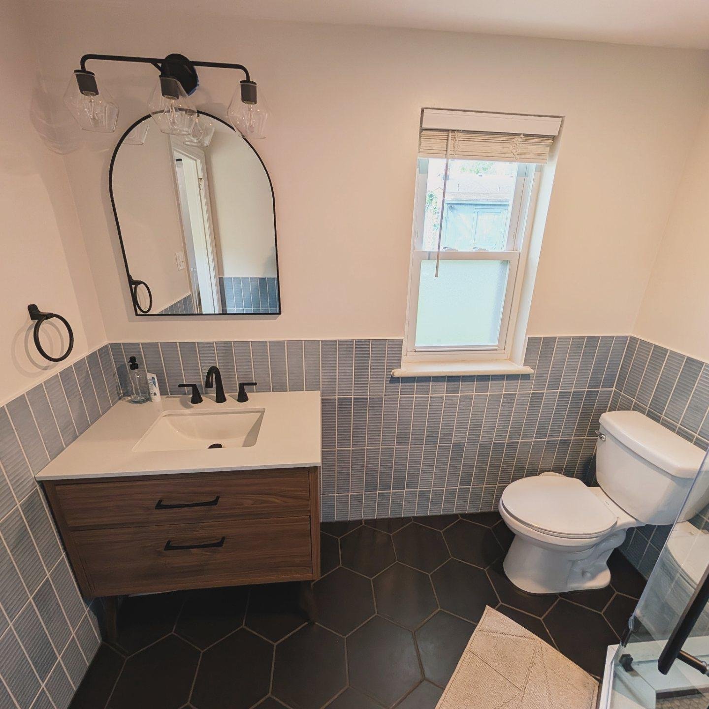 Close view of a walnut-look floating vanity with a white quartz top, black single-handle faucet, two black bar pulls, an arched black mirror, a three-bulb geometric black sconce, dusty blue vertical tile running as wainscoting, and large black hexagon floor tile.
