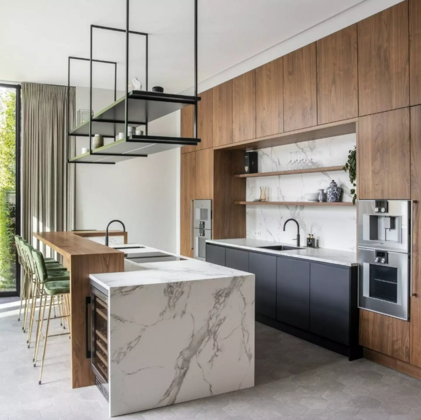 Modern open kitchen with walnut veneer tall cabinets, matte black lower cabinets, white marble waterfall island with built-in wine fridge, a raised wood bar extension holding cooktops, green velvet bar stools with gold legs, and a suspended black metal rectangular shelf fixture
