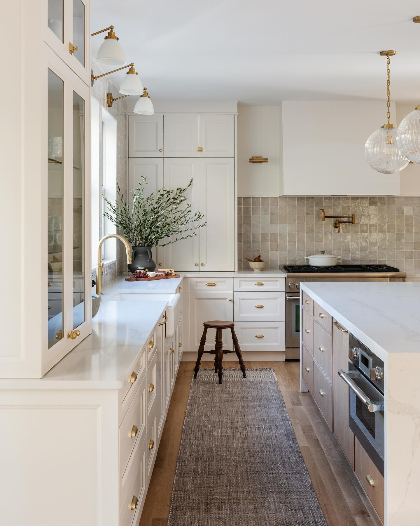 Cream shaker kitchen with warm earthen square tile backsplash, three brass swing-arm sconces above the sink, farmhouse sink with brass gooseneck faucet, olive branches in black vase, and whitewashed oak island with white quartz waterfall top