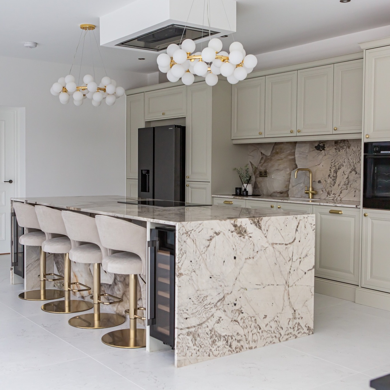 Dramatic brown and cream veined waterfall marble island with soft sage green cabinets, bubble chandelier, and velvet covered bar stools