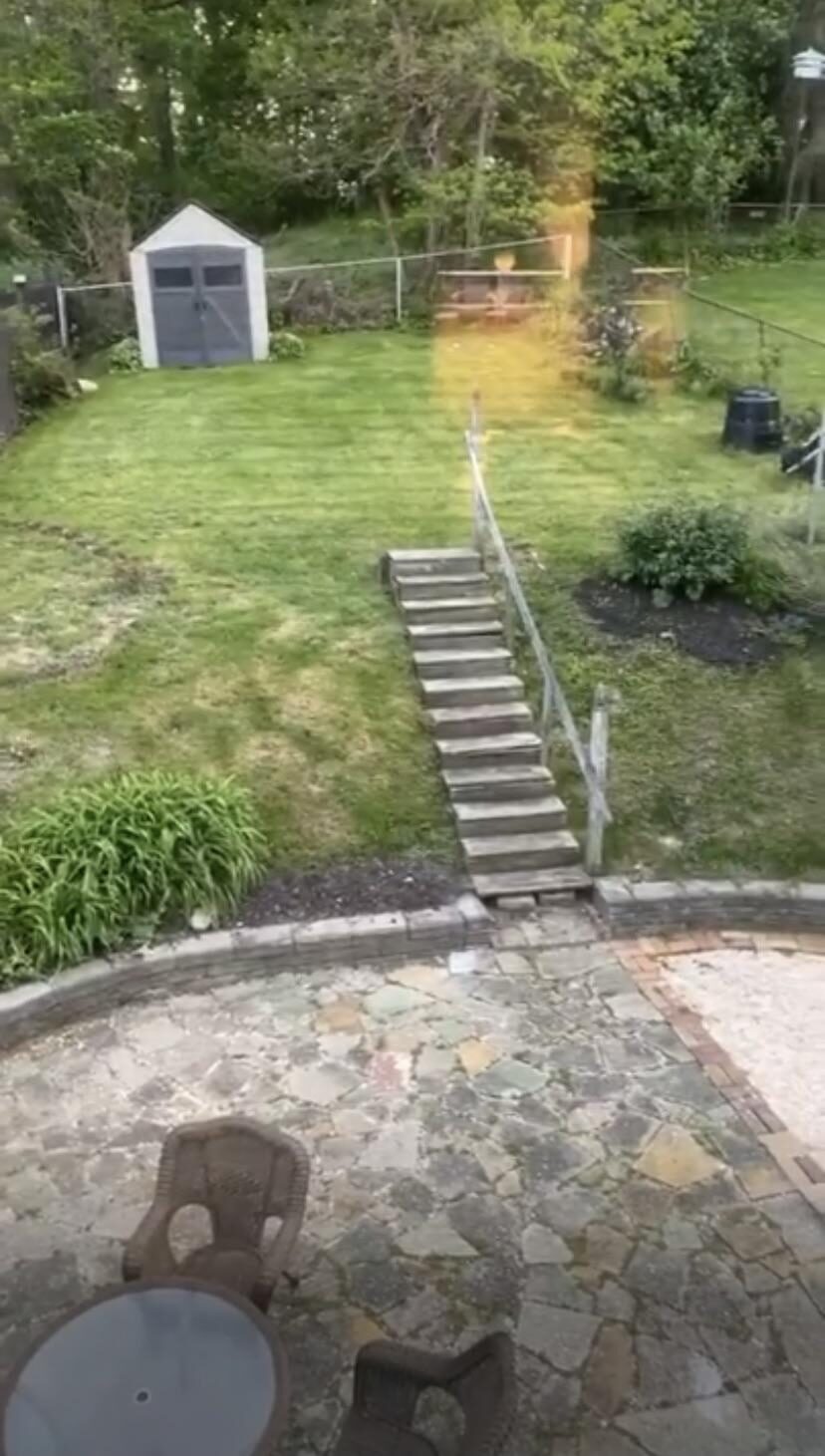 Overgrown backyard slope with weedy grass, a plastic shed at the top behind a chain link fence, a long run of worn wooden stairs, and a flagstone patio at the bottom.