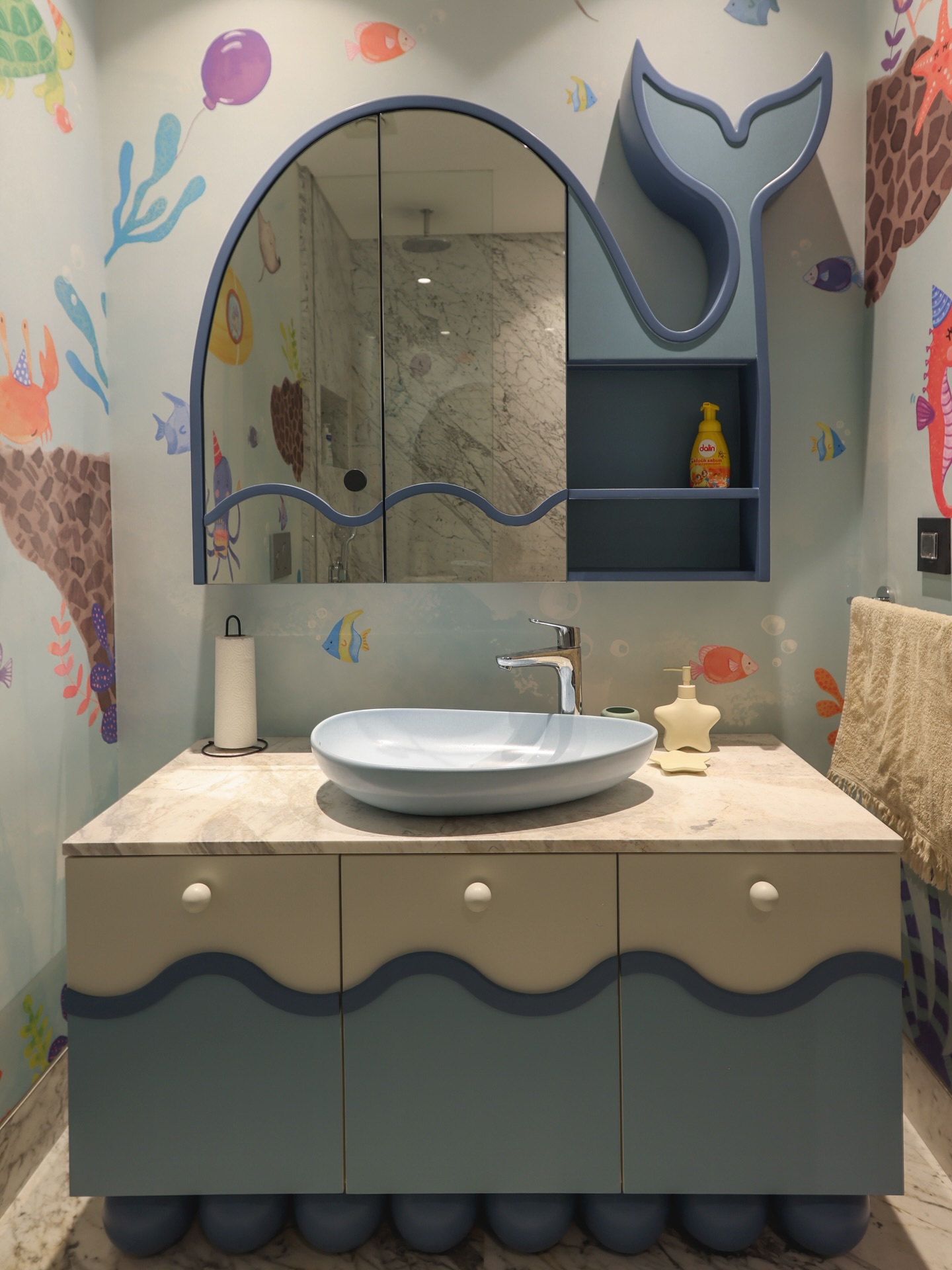 Blue whale tail cabinet beside an arched mirror above a wave-trimmed vanity with ocean mural wallpaper and oval blue vessel sink