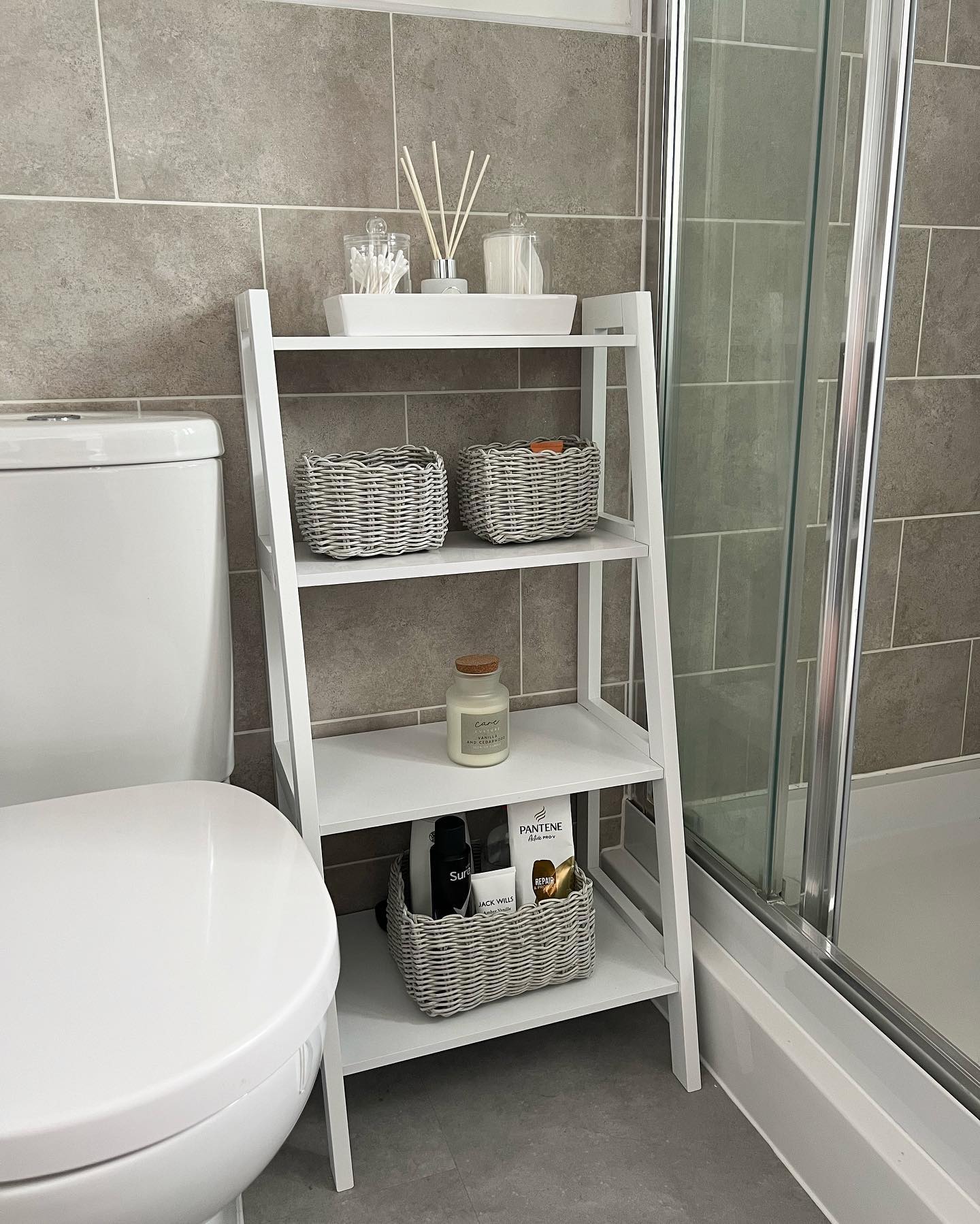 White four-tier angled ladder shelf standing in the narrow gap between a white toilet and glass shower door with gray stone tile walls, holding woven baskets and apothecary jars