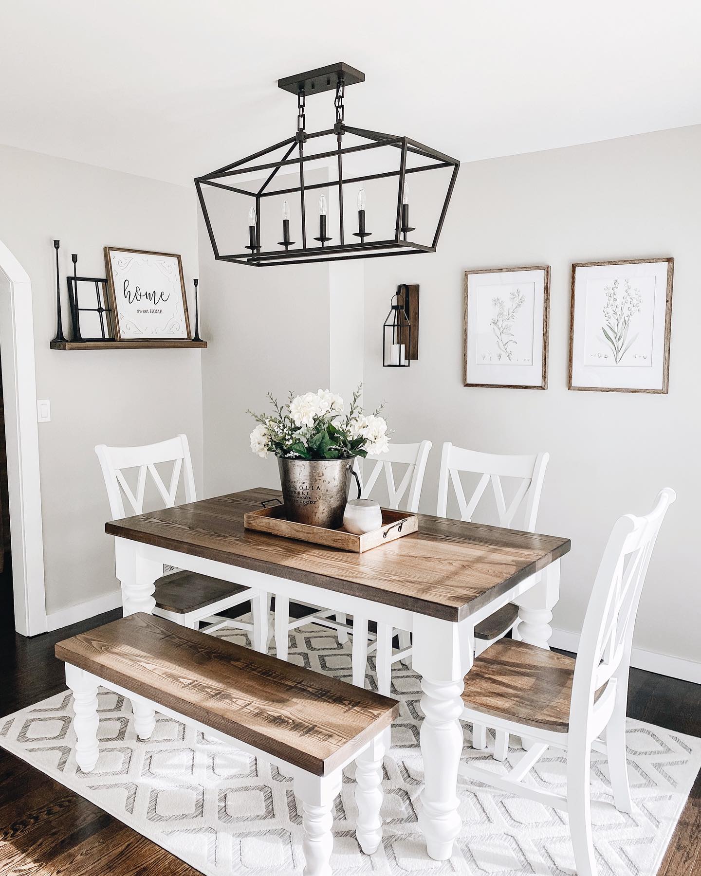 White farmhouse dining table with stained wood top, white X-back dining chairs, matching wood-topped bench on one side, large black cage lantern chandelier, and two framed botanical prints on the wall