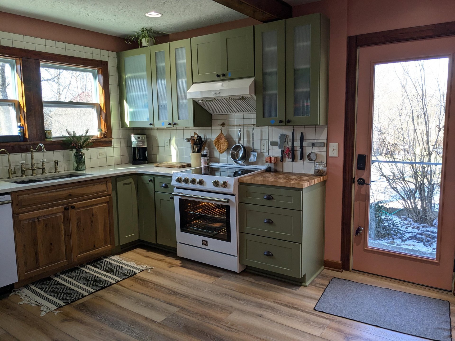 After view of the stove area with a white Forno range with brass knobs, butcher block counter, white square tile backsplash, and sage green cabinets with reeded glass uppers.