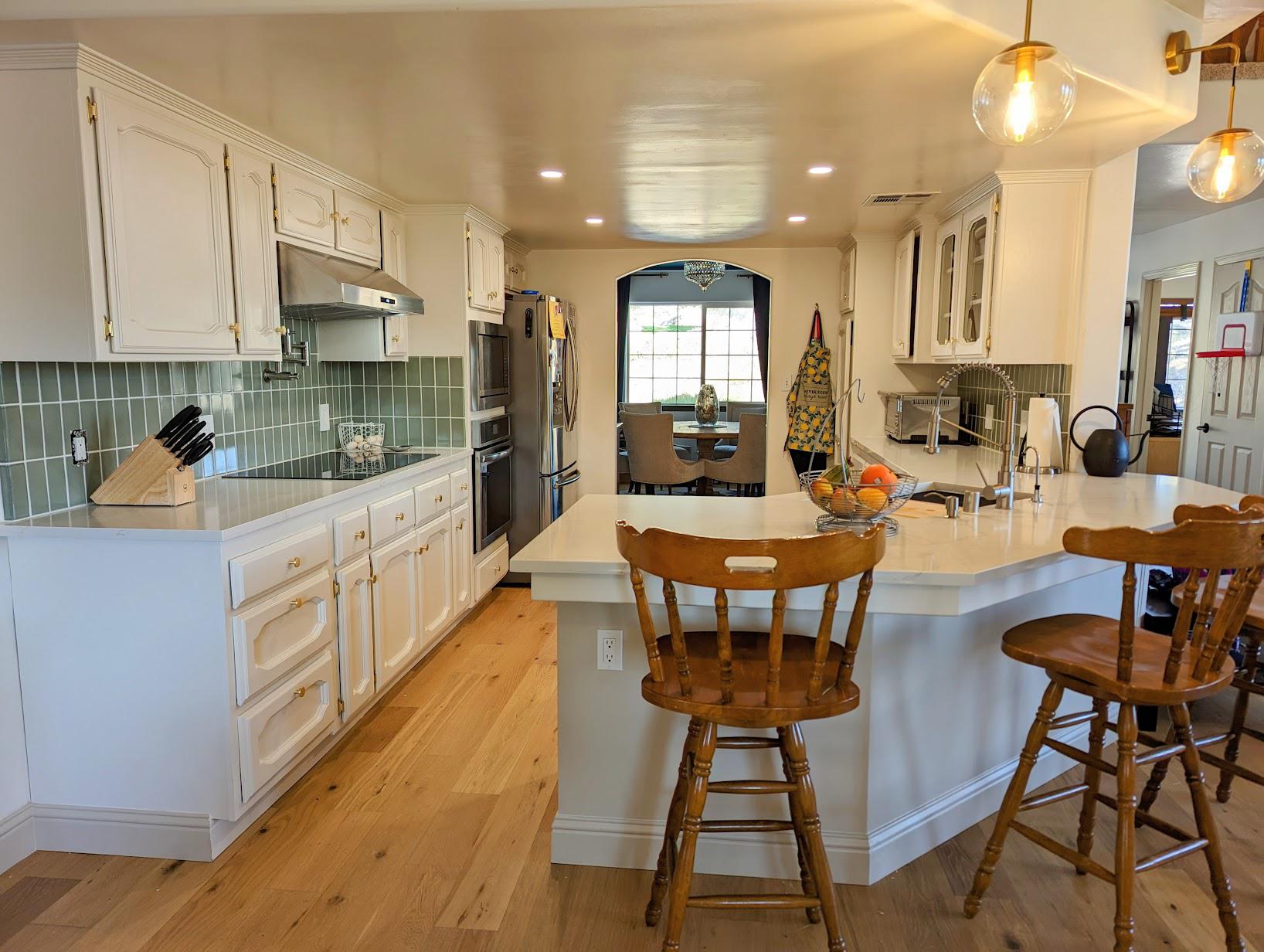 Side view of the renovated kitchen with white painted cabinets, sage green backsplash, a stacked wall oven and microwave, induction cooktop, and vintage wood counter stools pulled up to the quartz peninsula.