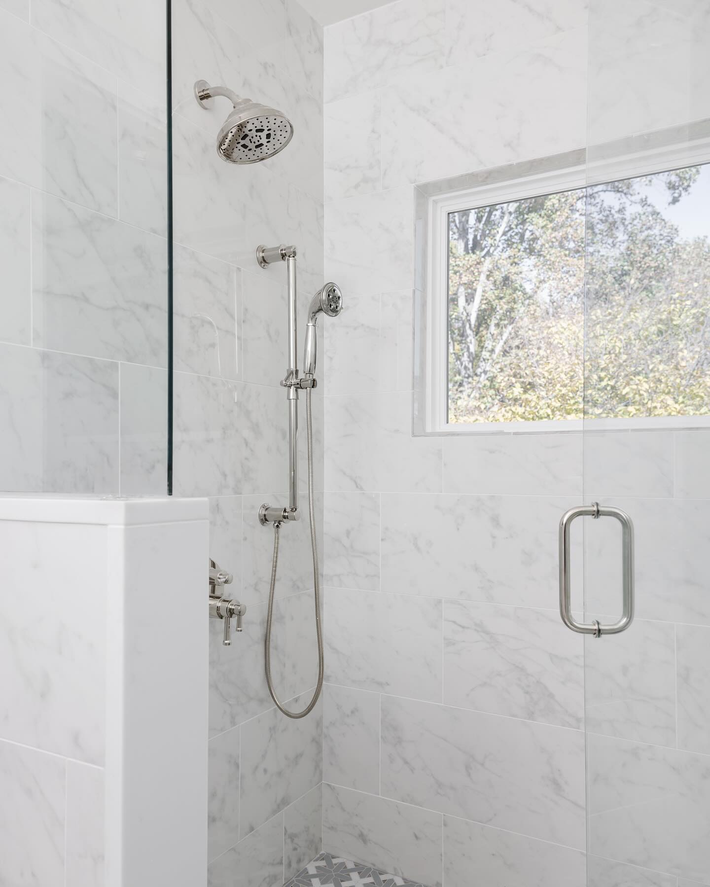 Clean white Carrara marble subway tile walk-in shower with polished nickel rainfall head, handheld on a slide bar, a window looking out to trees, and a glimpse of patterned floor tile