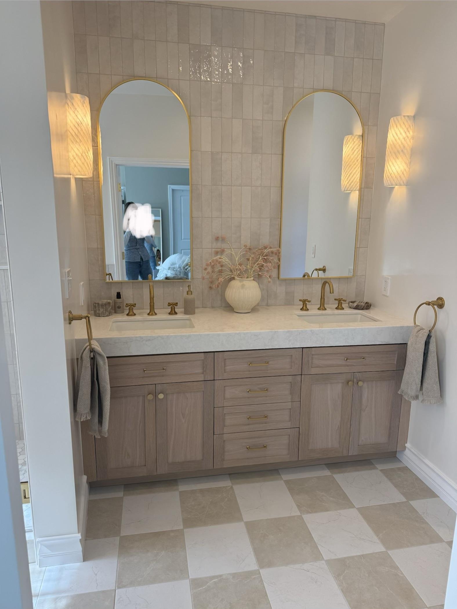 Light wood double vanity with white quartz counter, undermount sinks, brass cross-handle faucets, two tall arched brass mirrors, pleated plaster sconces, and a vertical oat zellige tile backsplash reaching the ceiling.