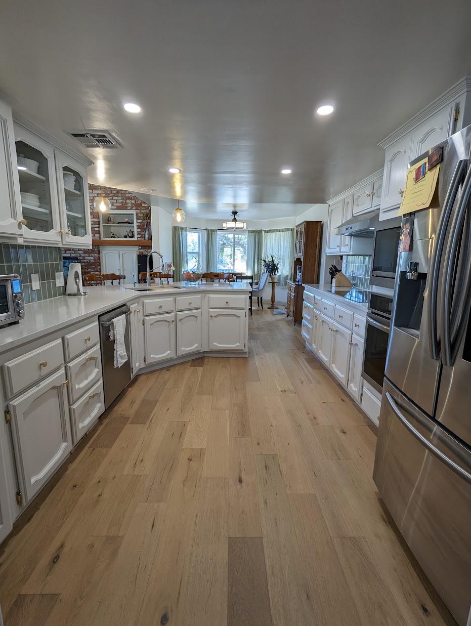 Full kitchen view after renovation showing white oak engineered flooring, white painted cabinets on both runs, sage backsplash, clear glass pendants hanging over the peninsula, stainless fridge, dishwasher, and the breakfast nook in the distance with the same brick accent wall.