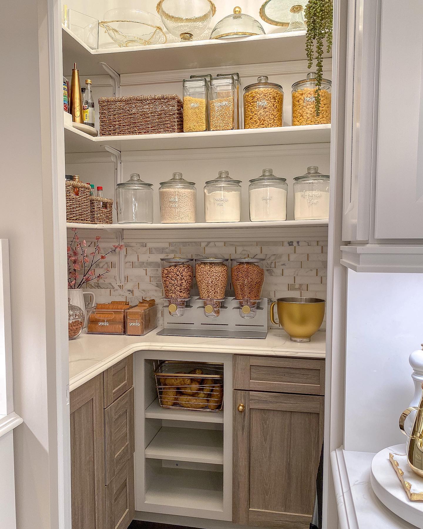 White pantry with floor-to-ceiling open shelves, labeled glass canisters for flour and sugar, three wall-mounted cereal dispensers, marble subway tile backsplash with gold inlay, and a gold stand mixer bowl on the counter