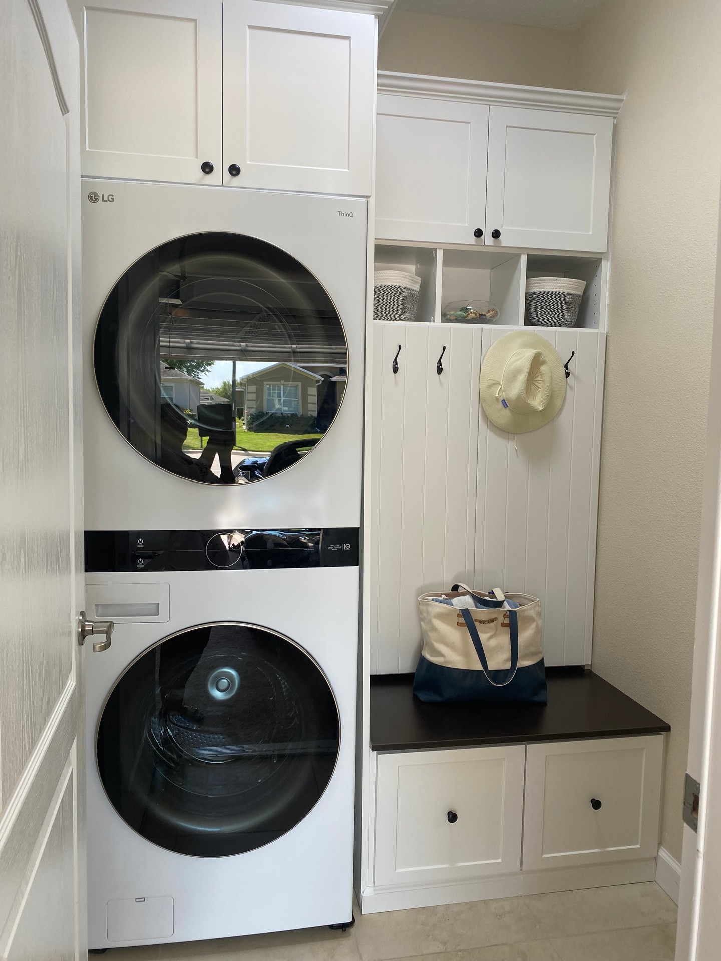 White shaker cabinets with black hardware, a stacked LG WashTower on the left, and a mudroom bench on the right with hooks holding a straw hat and canvas tote, a dark wood seat, and lower drawer storage over patterned tile