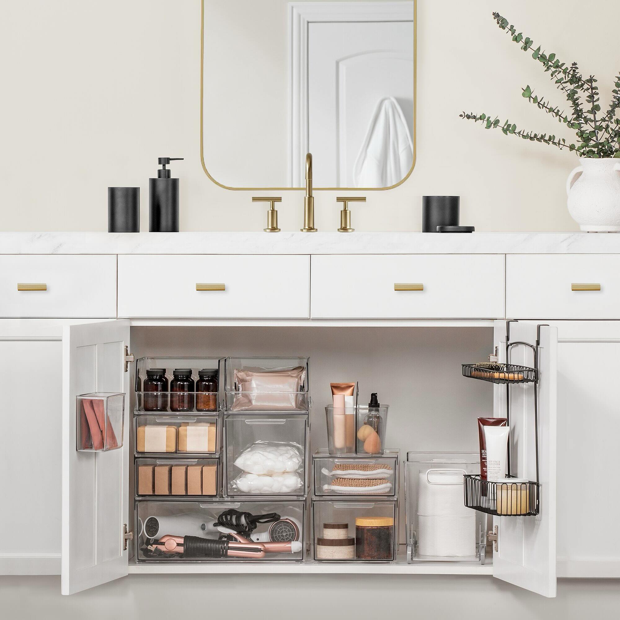 Open white bathroom vanity with multiple clear stackable storage bins holding amber pill bottles, cotton pads, soap bars, hair tools, and skincare, with a wire caddy on the door