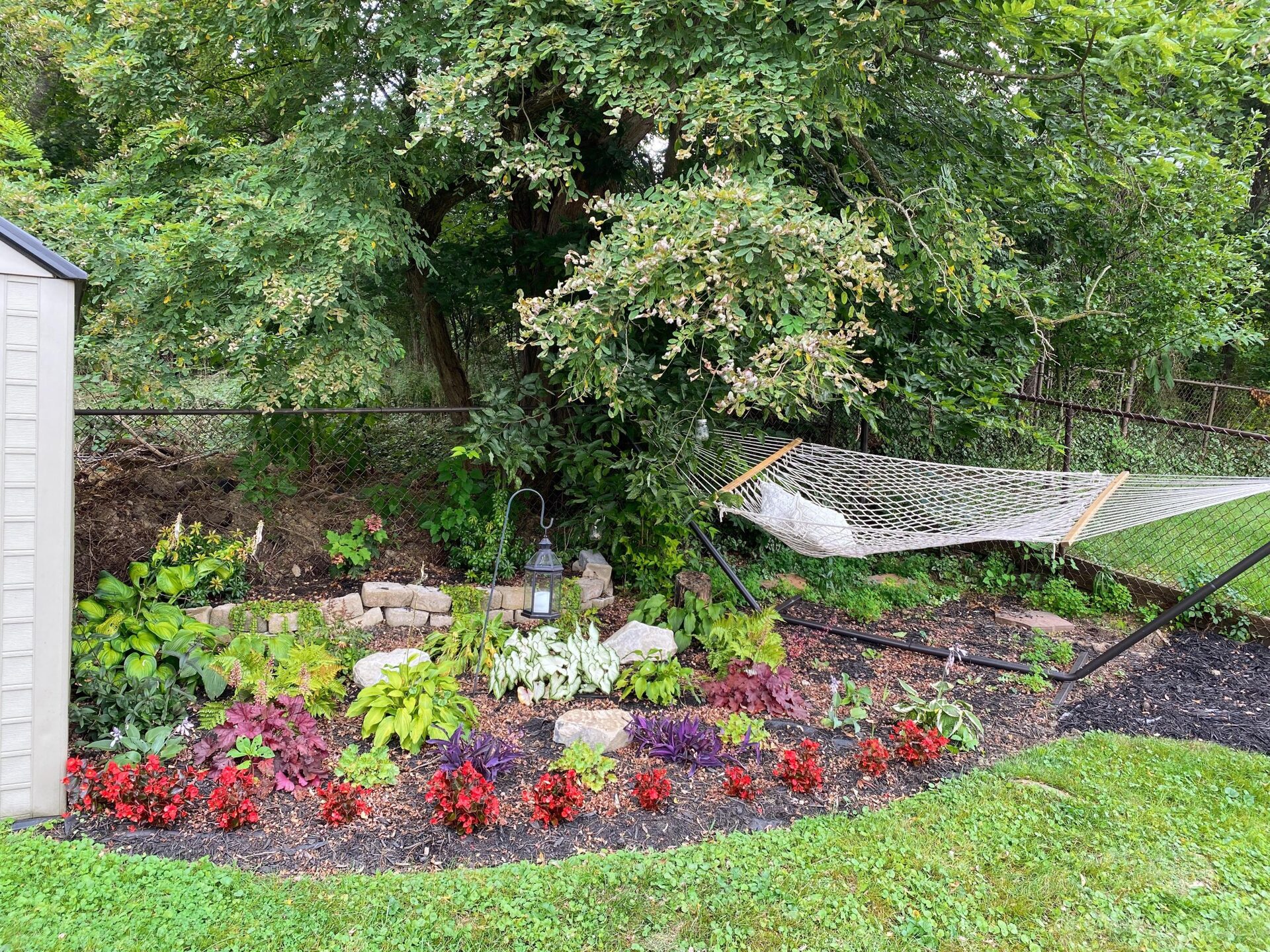 Round wicker dining table with four cushioned wicker chairs under a scalloped cream market umbrella, with climbing green vines up the wooden fence behind and a palm leaf in the foreground.