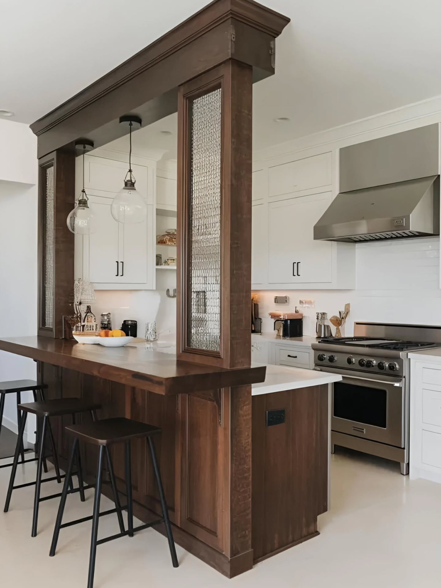 Open kitchen seen through a dark stained wood column frame with chicken-wire textured glass panels, white Shaker cabinets with black pulls, stainless gas range with vented hood, two clear glass globe pendants, and a solid dark wood bar top with simple black iron stools
