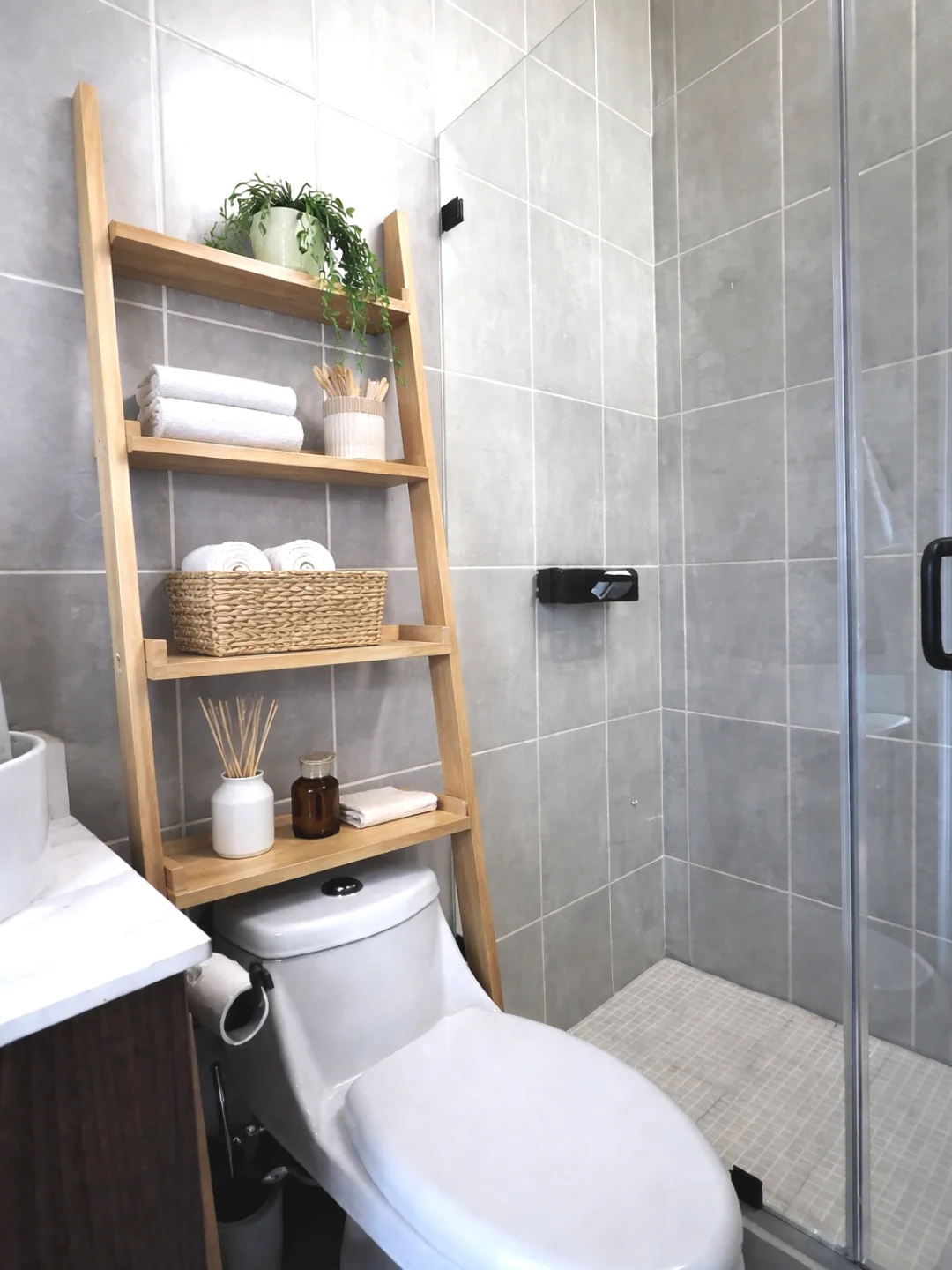 Natural wood leaning ladder shelf propped over the toilet with a trailing plant, rolled towels, a woven basket, and a reed diffuser in a gray tiled bathroom