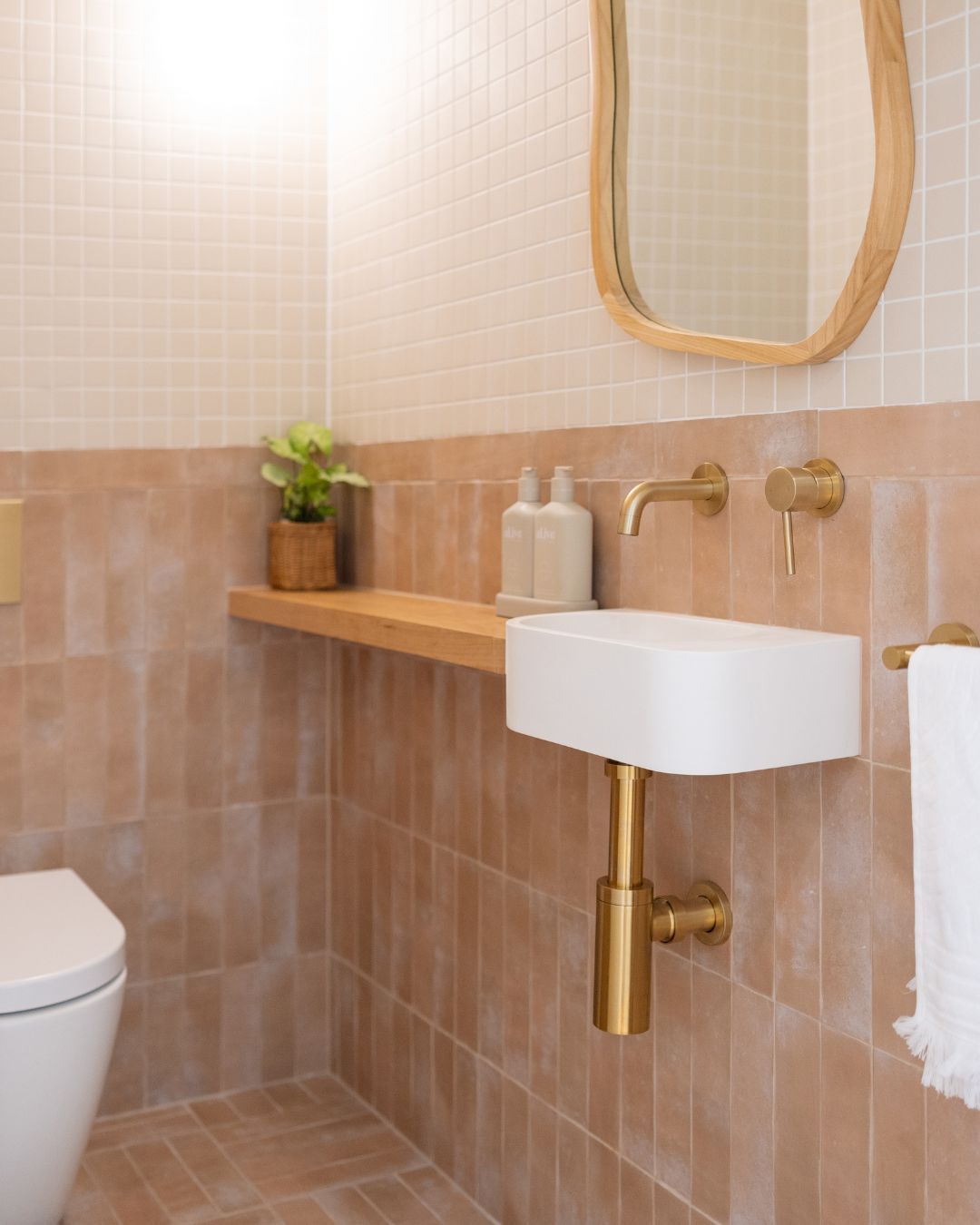Narrow wood ledge extending the counter beside a small white wall-mount sink with brass fixtures in a terracotta tiled powder room