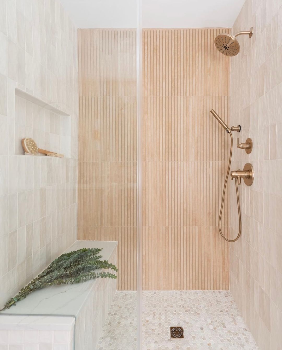 Spa-style walk-in shower with vertical fluted wood-look porcelain tile on the back wall, warm beige stone tile on the side walls, a marble bench with trailing eucalyptus, brass fixtures, and a penny-round hex floor