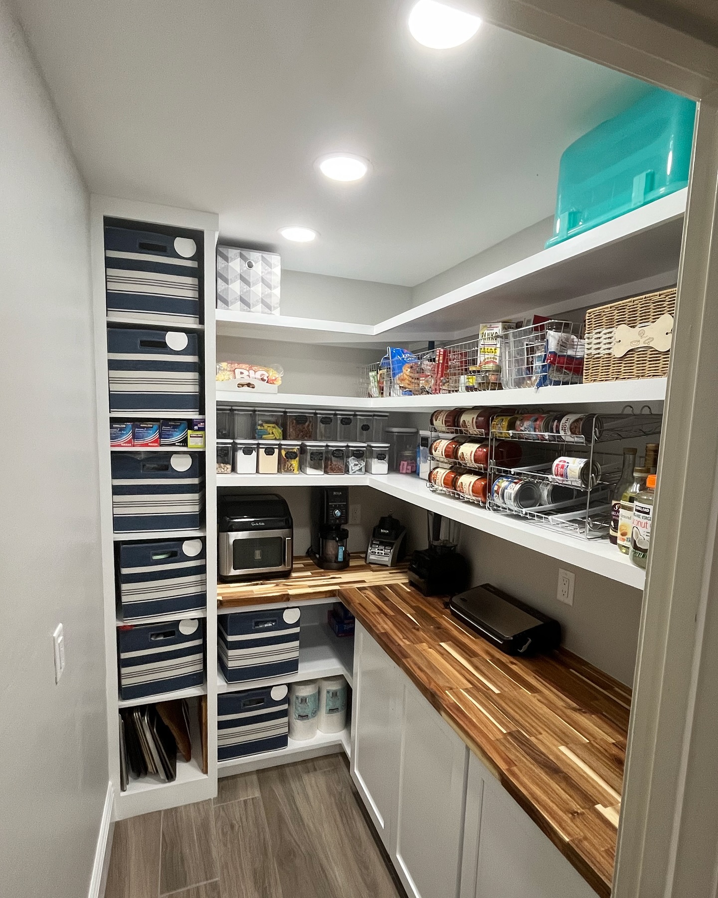 DIY walk-in pantry with raw pine floating shelves on black brackets, butcher block counters, two black cereal dispensers, four-slice toaster, air fryer, and gray plastic basket bins labeled with chalkboard tags
