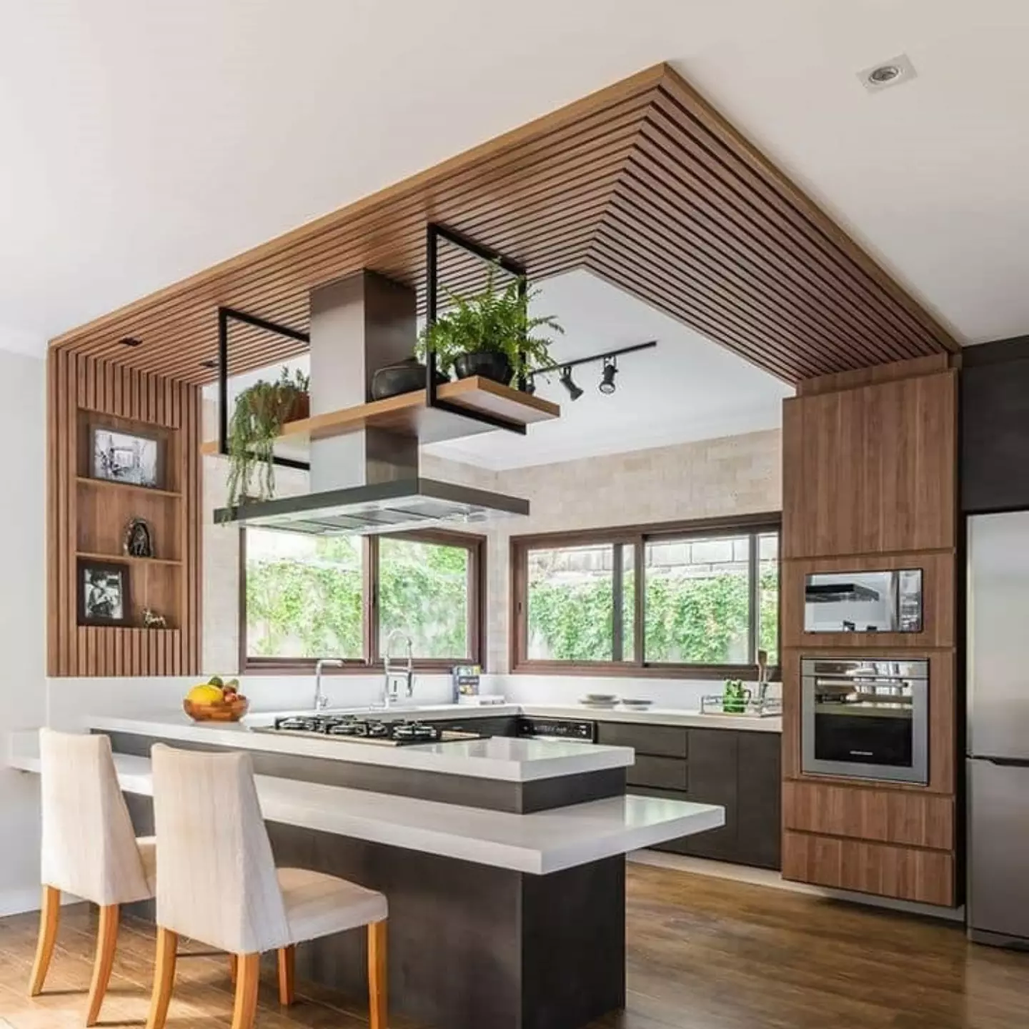 Open plan kitchen with a slatted wood ceiling panel wrapping down the side wall, white marble island with dark base and built-in cooktop, suspended black metal planter shelves holding ferns and greenery, large windows looking out at a vine-covered wall, and warm wood accent walls